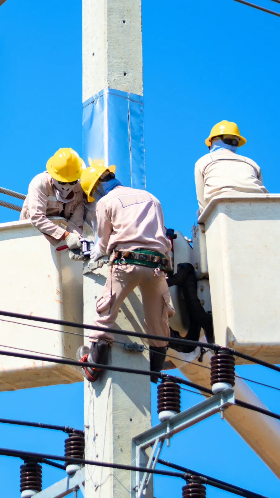 Equipe de eletricistas em manutenção no topo de poste de distribuição, com uniformes retardantes de chama, capacetes e cinturão de segurança