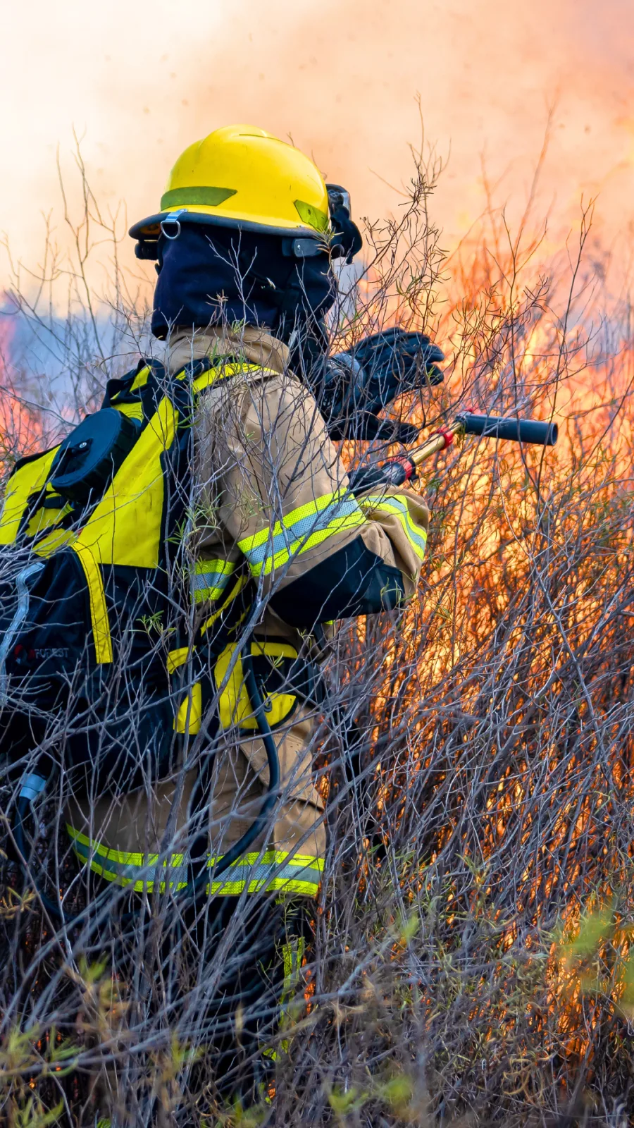 Brigadista florestal em combate a incêndio em vegetação densa, com jaqueta retardante de chama, faixas retrorrefletivas, capacete e balaclava
