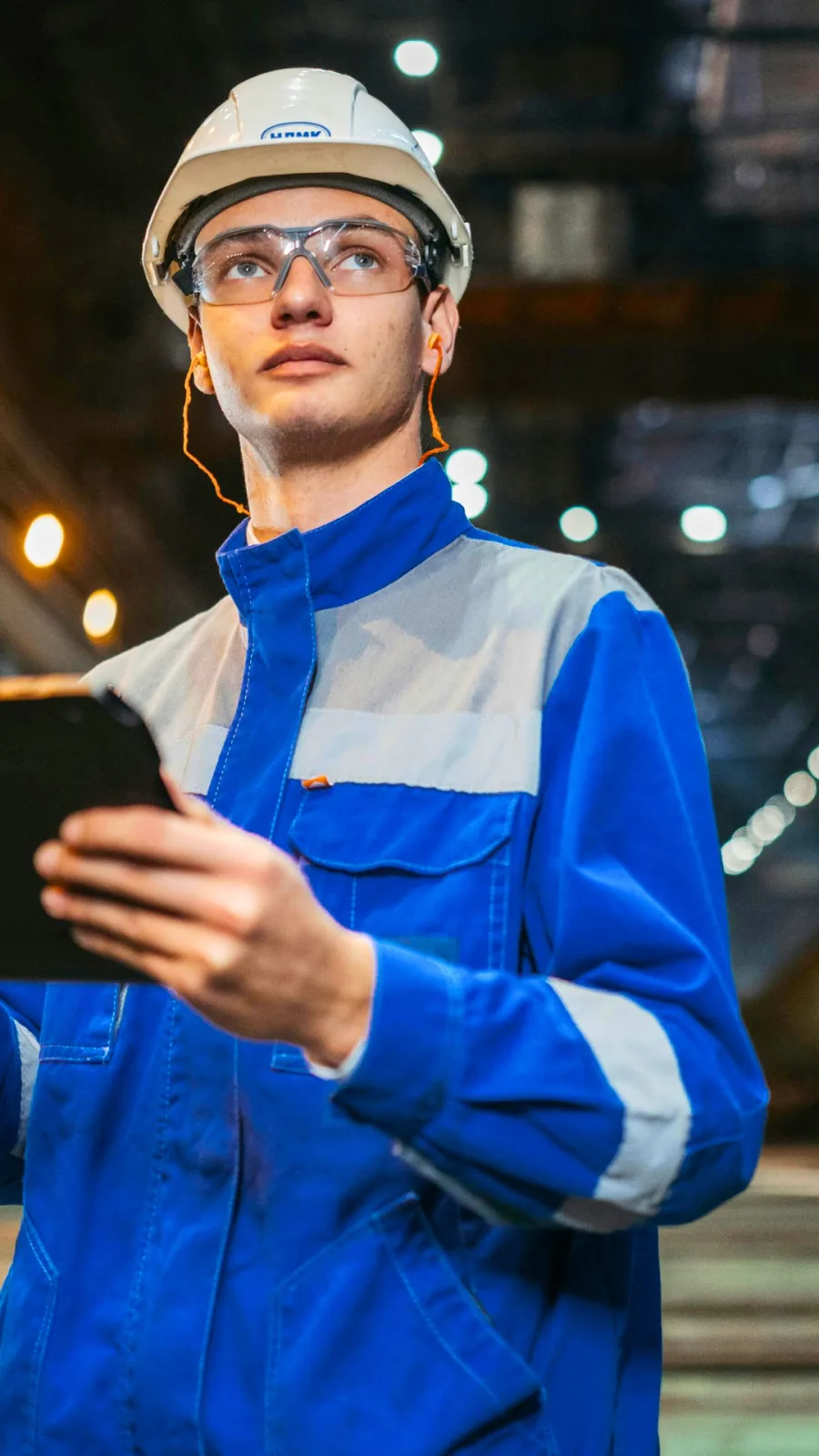 Profissional com jaqueta de uniforme industrial azul e detalhes refletivos, capacete e óculos de proteção, consultando tablet em ambiente fabril